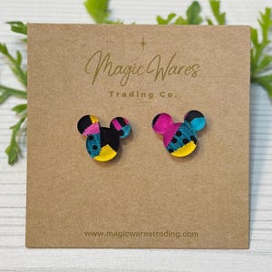 May include: A pair of Mickey Mouse-shaped stud earrings. Each earring features a patchwork design with black, yellow, pink, and teal colors. The earrings are displayed on a brown card with the text "Magic Wares Trading Co." and the website address.