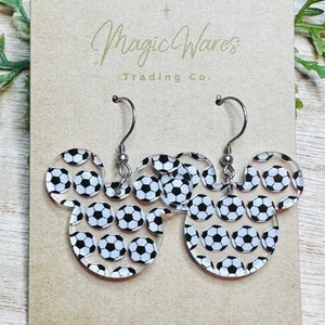 May include: A pair of clear acrylic earrings shaped like Mickey Mouse ears with a soccer ball pattern. The earrings are on a brown card with the text "Magic Wares Trading Co."
