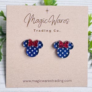 May include: A pair of blue and white star-patterned mouse-shaped earrings with red glitter bows. The earrings are displayed on a tan card with the text "Magic Wares Trading Co." and the website address.