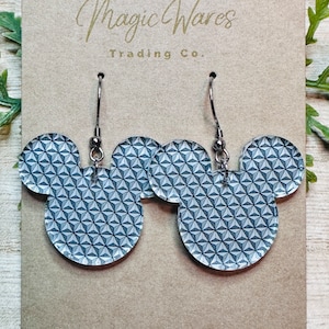 May include: A pair of silver dangle earrings with a blue and white geometric pattern in the shape of Mickey Mouse heads. The earrings are on a brown card with the text "Magic Wares Trading Co."
