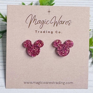 May include: Pink glitter Mickey Mouse stud earrings on a light pink card. The card has the text "Magic Wares Trading Co." and the website address www.magicwarestrading.com.