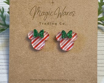 May include: A pair of Minnie Mouse-shaped earrings with red and white striped detailing. Each earring features a green glitter bow. The earrings are displayed on a brown card with the text "Magic Wares Trading Co."