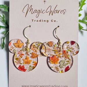May include: A pair of clear resin earrings shaped like mouse heads, featuring embedded autumn leaves and gold glitter. The earrings hang from gold-toned metal hooks and are displayed on a beige card labeled Magic Wares Trading Co.
