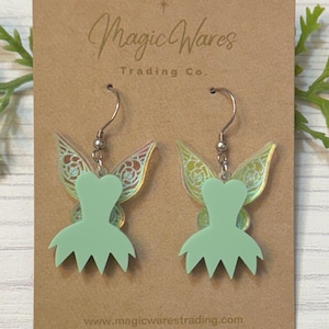 May include: Mint green fairy earrings with iridescent wings and a dress-shaped pendant. The earrings hang from silver-colored hooks. The earrings are displayed on a brown card with the text "Magic Wares Trading Co." and the website address.