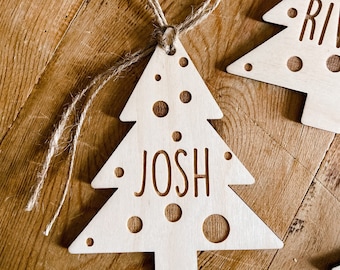 Könnte beinhalten: Ein Weihnachtsbaum-Anhänger aus Holz mit dem eingravierten Namen "Josh". Der Anhänger ist hellbraun und hat eine Schlaufe aus Schnur zum Aufhängen.