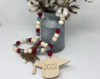 May include: A graduation cap ornament with the year 2022 engraved on it. The ornament is made of wood and has a tassel. The ornament is hanging from a beaded garland with red and gray beads.