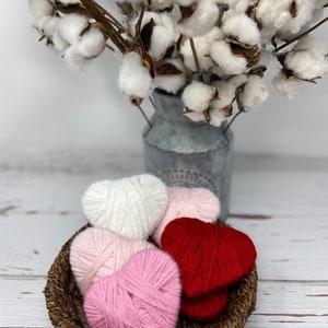 May include: A wicker basket filled with yarn hearts in pink, white, and red. The basket is sitting on a white wooden surface.