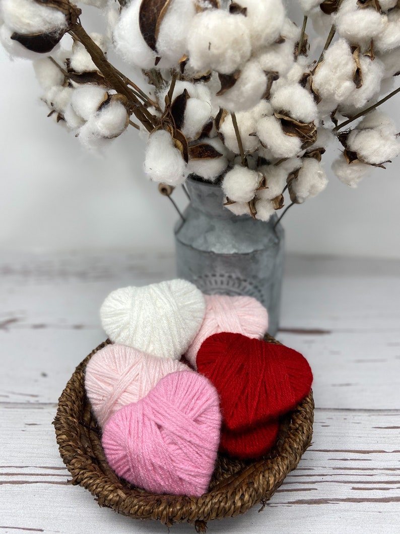 May include: A wicker basket filled with yarn hearts in pink, white, and red. The basket is sitting on a white wooden surface.