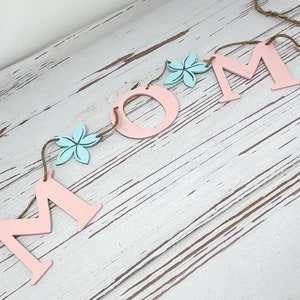 May include: A pink and blue wooden banner with the word "MOM" spelled out in large letters. The letters are strung together with twine and decorated with small wooden flowers in yellow, blue, and pink.