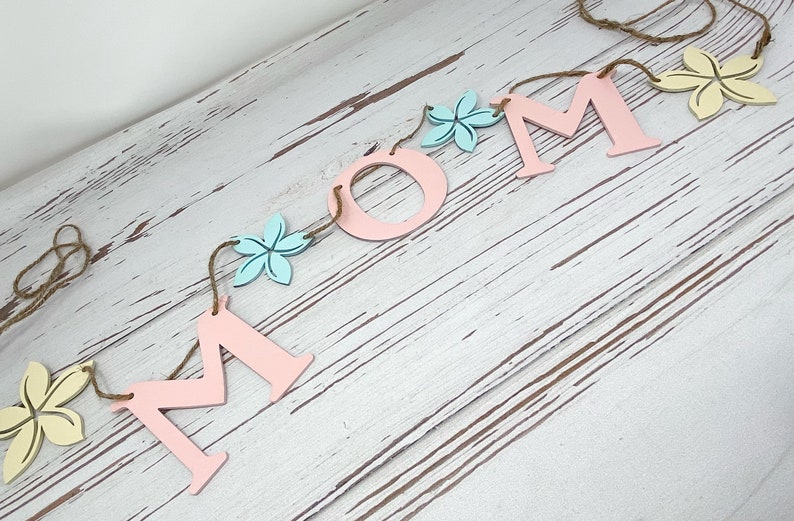 May include: A pink and blue wooden banner with the word "MOM" spelled out in large letters. The letters are strung together with twine and decorated with small wooden flowers in yellow, blue, and pink.