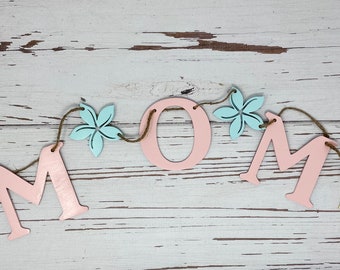 May include: A pink wooden MOM banner with yellow and blue flowers strung on twine. The banner is on a white wood background.