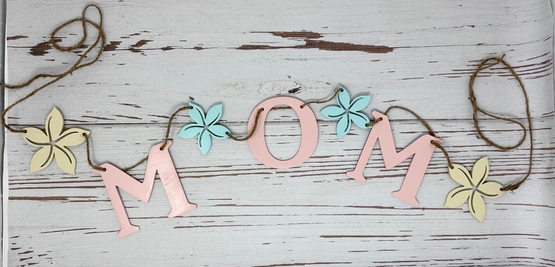 May include: A pink wooden MOM banner with yellow and blue flowers strung on twine. The banner is on a white wood background.