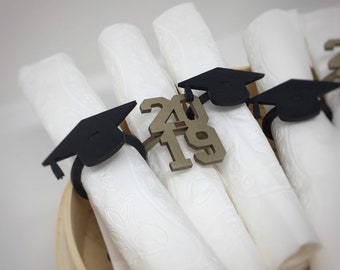 Puede incluir: Anillos de servilleta de graduación dorados con el año 2019 en ellos. Los anillos son negros y dorados y tienen un gorro de graduación en la parte superior.