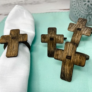 May include: Four wooden cross napkin rings in a dark brown wood stain. The rings are on a light blue tablecloth. One ring is on a white napkin.