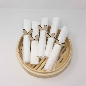 Puede incluir: Un juego de seis servilletas de tela blancas con anillos de servilleta de madera con forma de conejo. Los anillos son de color marr&oacute;n claro y tienen un dise&ntilde;o sencillo. Las servilletas est&aacute;n dispuestas en una vaporera de bamb&uacute;.