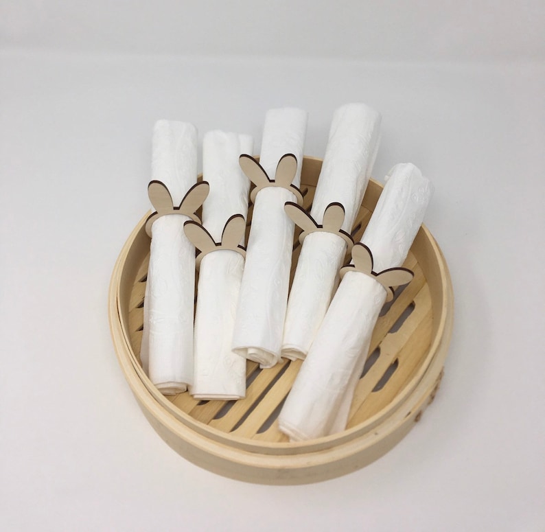 Puede incluir: Un juego de seis servilletas de tela blancas con anillos de servilleta de madera con forma de conejo. Los anillos son de color marr&oacute;n claro y tienen un dise&ntilde;o sencillo. Las servilletas est&aacute;n dispuestas en una vaporera de bamb&uacute;.
