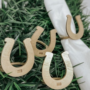 May include: Four gold horseshoe napkin rings on a green grass background. The rings are made of wood and have a rustic look.