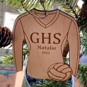 May include: Wooden ornament shaped like a volleyball jersey. The jersey is light brown with the letters "GHS", the name "Natalie", and the year "2022" engraved on it. A volleyball is at the bottom. The ornament hangs from a twine loop.