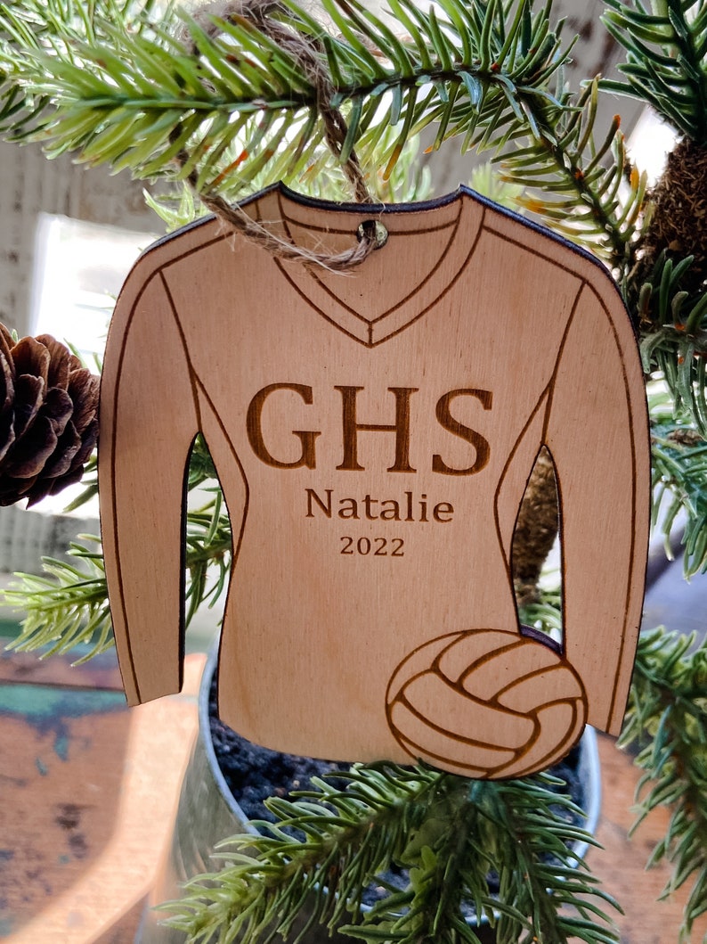 May include: Wooden ornament shaped like a volleyball jersey. The jersey is light brown with the letters "GHS", the name "Natalie", and the year "2022" engraved on it. A volleyball is at the bottom. The ornament hangs from a twine loop.