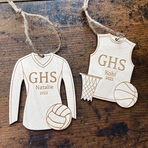May include: Two wooden sports ornaments with twine hangers. One is a volleyball jersey with "GHS", "Natalie", and "2022". The other is a basketball jersey with "GHS", "Kobi", and "2022". A volleyball and basketball are also included.
