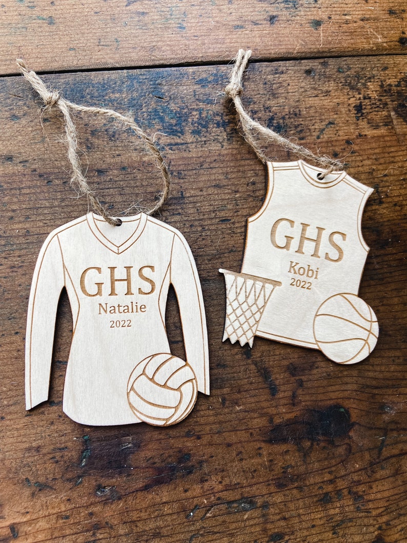 May include: Two wooden sports ornaments with twine hangers. One is a volleyball jersey with "GHS", "Natalie", and "2022". The other is a basketball jersey with "GHS", "Kobi", and "2022". A volleyball and basketball are also included.