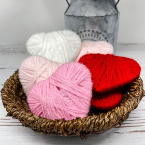 May include: Four yarn hearts in a wicker basket. The hearts are white, pink, and red.