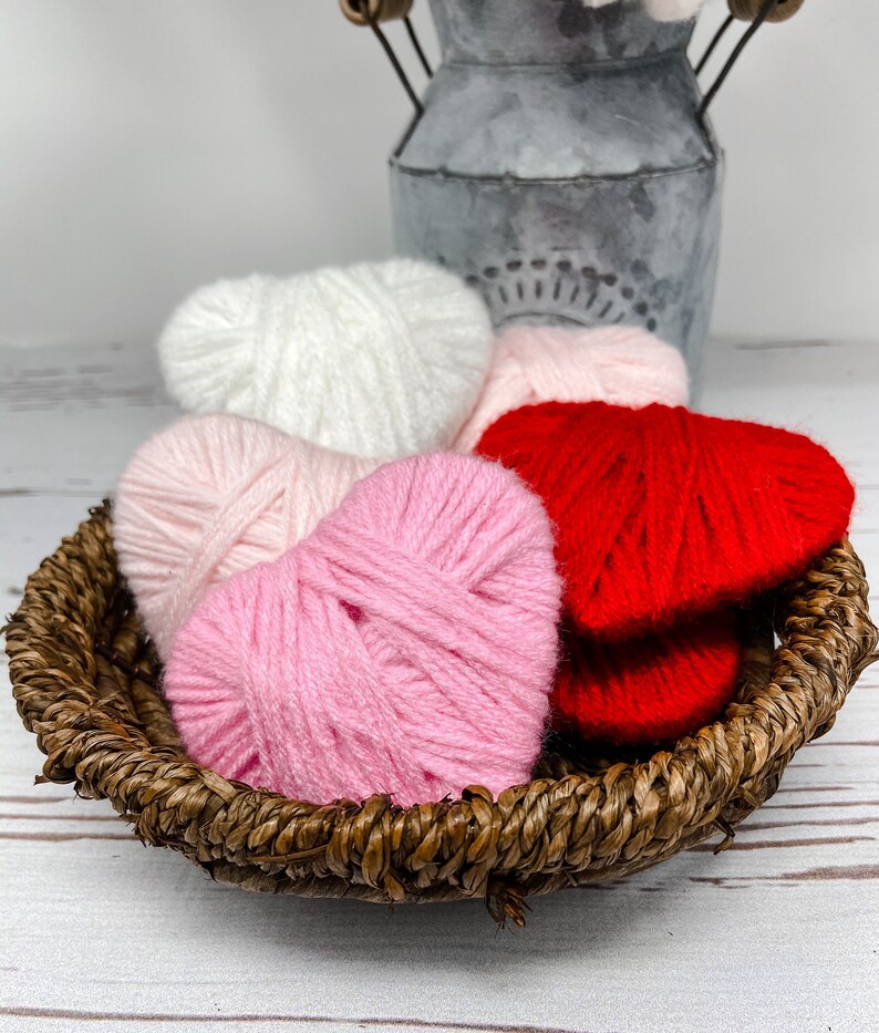 May include: Four yarn hearts in a wicker basket. The hearts are white, pink, and red.