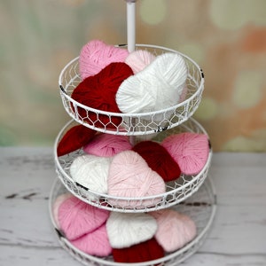 May include: A white tiered stand with three levels, each holding yarn hearts in pink, red, and white. The stand is on a white wooden surface.