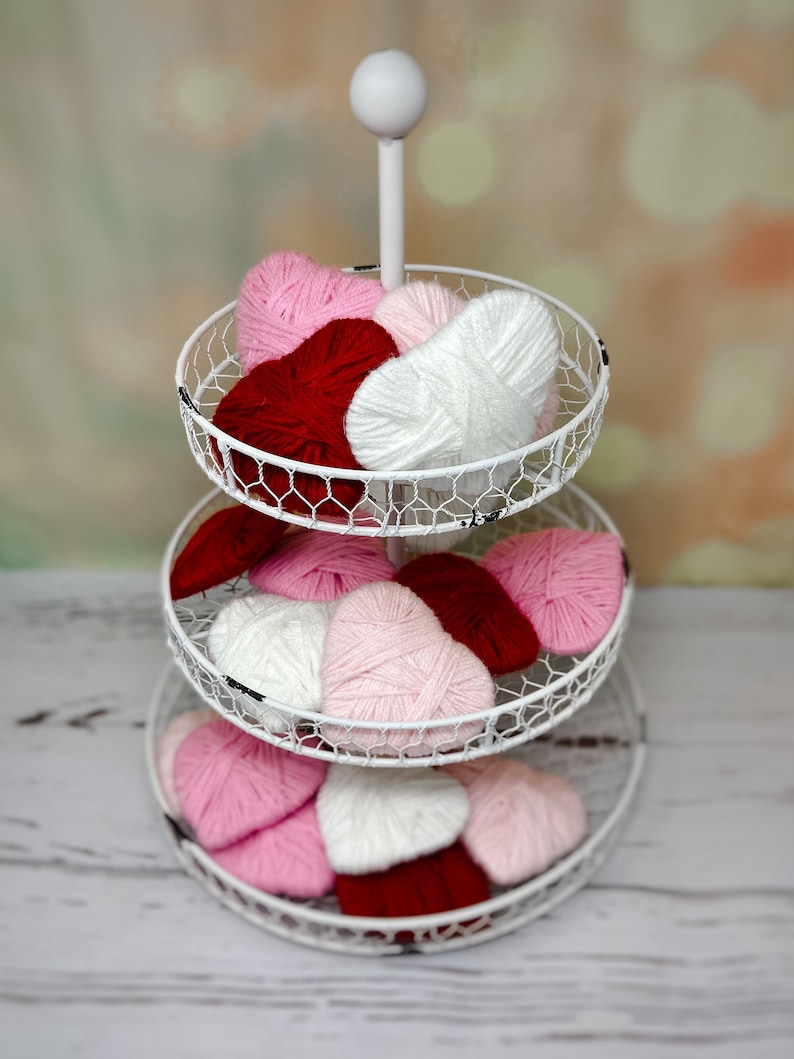 May include: A white tiered stand with three levels, each holding yarn hearts in pink, red, and white. The stand is on a white wooden surface.