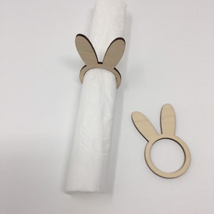 Puede incluir: Dos anillos de servilleta de madera con forma de conejo. Un anillo sostiene una servilleta blanca. El otro anillo est&aacute; colocado plano sobre una superficie blanca.