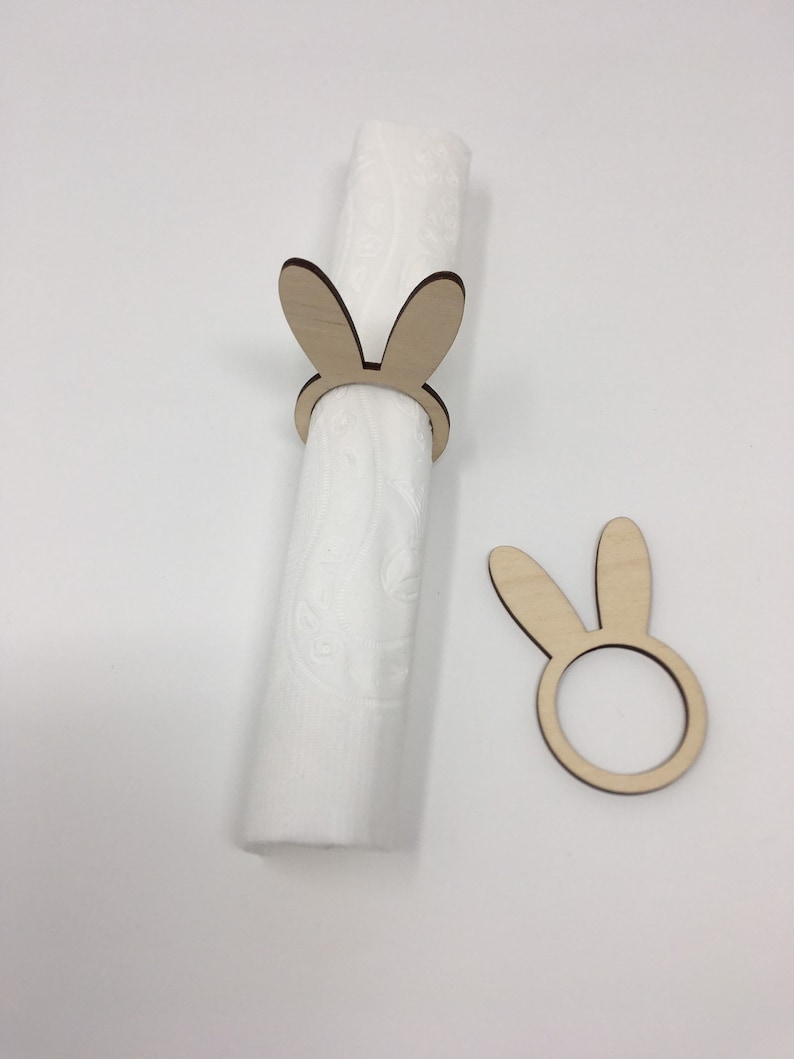 Puede incluir: Dos anillos de servilleta de madera con forma de conejo. Un anillo sostiene una servilleta blanca. El otro anillo est&aacute; colocado plano sobre una superficie blanca.