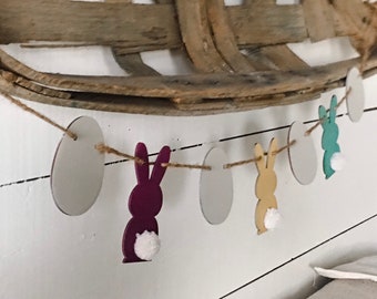 May include: Easter-themed banner featuring wooden egg and bunny cutouts. The banner includes white egg shapes and colorful bunnies in purple, yellow, and teal, each with a fluffy white tail. Strung on twine, the banner is set against a white background.