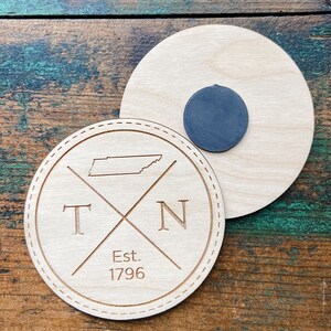 May include: Wooden coaster with a brown wood finish. The coaster features a silhouette of the state of Tennessee with the letters "TN" and "Est. 1796" engraved on it.