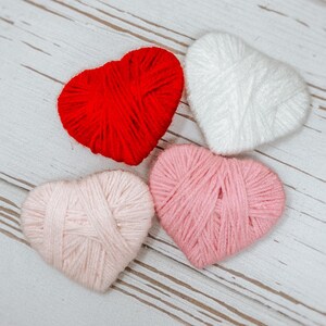 May include: Four yarn hearts in different colors: red, white, pink, and light pink. The hearts are arranged in a circle on a white wooden surface.