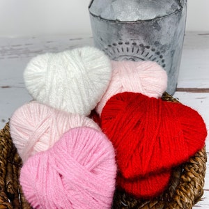 May include: Four yarn hearts in a wicker basket. The hearts are white, pink, and red.