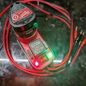 May include: A red and black Milwaukee True Mode battery charger with a green and red light indicator. The charger is connected to a red and black cable.