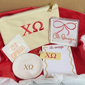 May include: A collection of Chi Omega sorority merchandise, including a canvas pouch with red embroidered letters, a clear acrylic frame with a pink bow design, a small dish, a keychain, and a notepad. The items are arranged on red tissue paper.