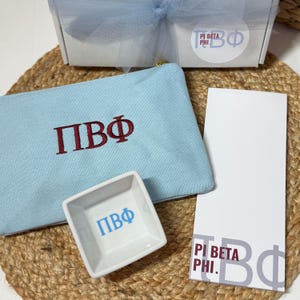 May include: A light blue zippered pouch and a small white square dish, both embroidered with "ΠΒΦ" in maroon and blue. A white box with a blue bow and a white notepad with "PI BETA PHI" are also visible. The items are arranged on a woven mat.
