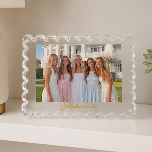 May include: A clear acrylic rectangular photo frame with a decorative scalloped edge. The frame holds a photograph of five individuals standing in front of a white building. The text Alpha Phi is printed in gold script at the bottom center.