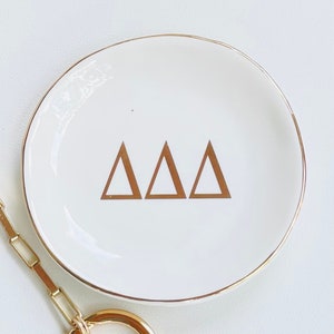 May include: White ceramic trinket dish with a gold rim and three gold triangles in the center. The triangles are arranged in a row and have the letters "ΔΔΔ" inside.