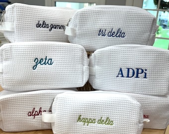 May include: A collection of white waffle-textured cosmetic bags with different sorority names embroidered on them. The names include: Zeta, Tri Delta, ADPi, Kappa Delta, Delta Gamma, and Alpha.