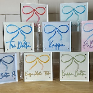 May include: Seven acrylic blocks with watercolor bow designs and sorority names. The blocks are stacked in a pyramid shape. The sorority names are: Tri Delta, Kappa, Pi Beta, Delta Pi, Kappa Alpha Theta, and Kappa Delta.