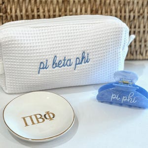 May include: A white waffle-textured cosmetic bag embroidered with "pi beta phi" in blue script. A small, round, white dish with gold trim and "ΠΒΦ" in gold lettering. A blue marbled hair claw clip with "pi phi" in white script.
