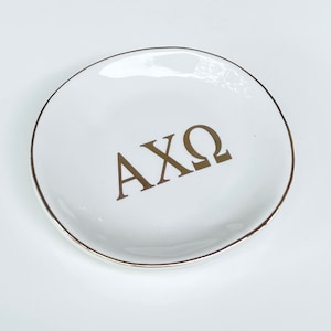 May include: White ceramic trinket dish with a gold rim and the letters AXQ in gold lettering.