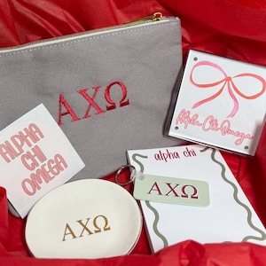 May include: A collection of Alpha Chi Omega merchandise, including a gray zippered pouch with red embroidered Greek letters, a white notepad, a small dish, a keychain, and a sticker. The items are arranged in a red gift box.