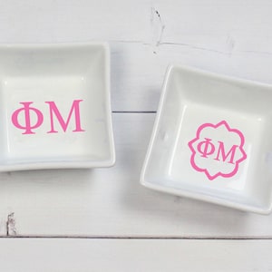 May include: Two white ceramic ring dishes with pink lettering. One dish has the letters "ΦΜ" in a simple font. The other dish has the letters "ΦΜ" in a decorative font inside a decorative frame.