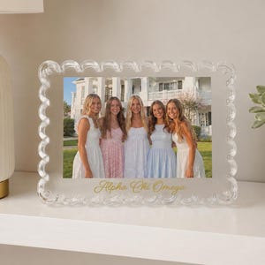 May include: A clear acrylic picture frame with a scalloped edge design. The frame holds a photograph of five individuals standing in front of a white building. The text Alpha Chi Omega is printed in gold script at the bottom of the frame.