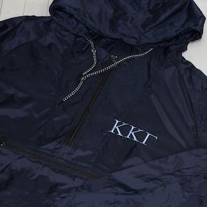 May include: A navy blue hooded windbreaker with a black zipper and a white drawstring. The jacket has a pocket on the left side and the letters "KKT" embroidered on the front in light blue.