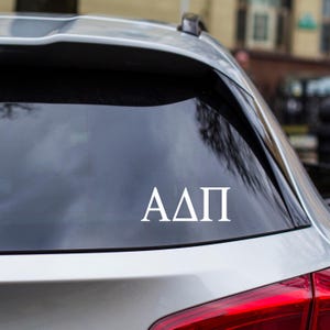May include: White vinyl decal with the Greek letters ΑΔΠ, applied to the rear window of a vehicle. The letters are in a sans-serif font and are centered on the glass. The car's red taillight is visible.