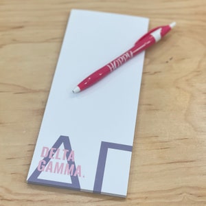 May include: A white notepad with the text "Delta Gamma" in pink and purple, and a pink pen with the word "Happy" printed on it. The notepad and pen are on a light wood surface.